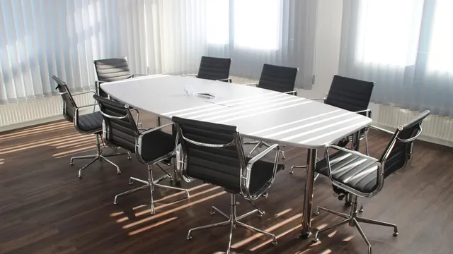 Une table de réunion blanche entourée de chaises vides, dans une salle baignée de lumière.