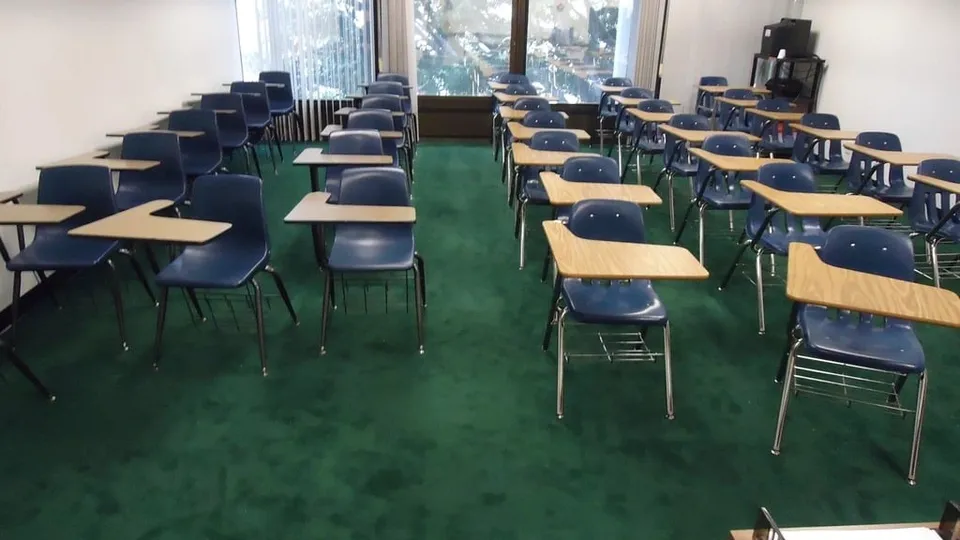 Une salle de classe vide : des rangées de tables et de chaises sur une moquette verte.